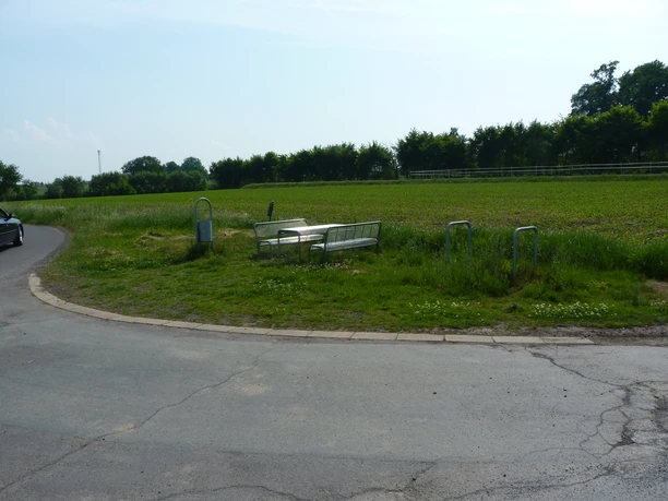 Eine ruhige Landstraße mit einer Bank und Tisch inmitten grüner Wiesen und umgeben von Bäumen.