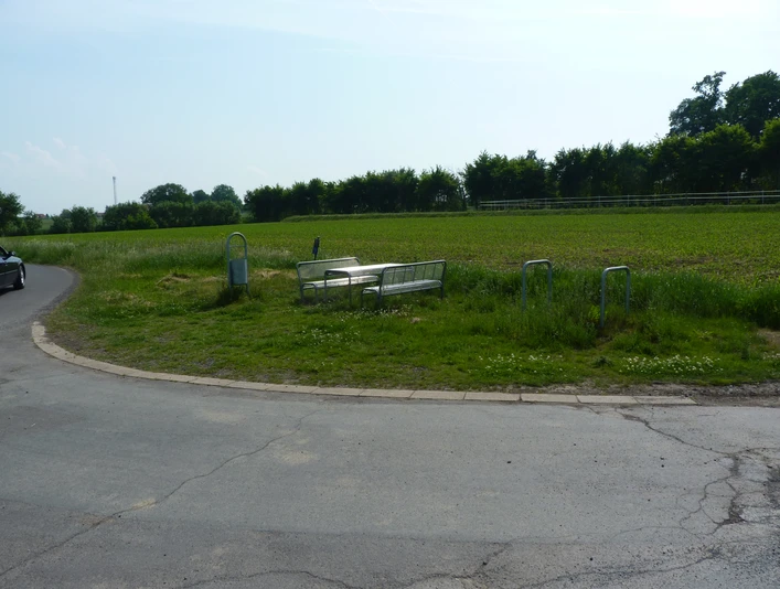 Eine ruhige Landstraße mit einer Bank und Tisch inmitten grüner Wiesen und umgeben von Bäumen.