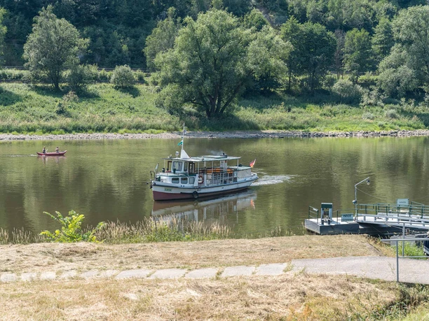 Fähre Schmilka Ein kleines Boot fährt auf einem ruhigen Fluss, während im Hintergrund ein Kanu mit zwei Personen paddelt; umgeben von grüner Landschaft.
