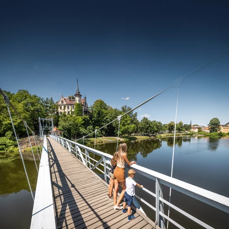 Mother and child on a suspension bridge over a river, castle in the background.