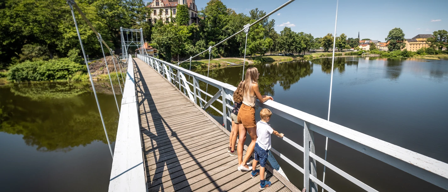 grimma-haengebruecke-gattersburg-philipp-herfort-photography-2042.jpg-2042.jpg Mutter und Kind auf einer Hängebrücke über einem Fluss, Schloss im Hintergrund.