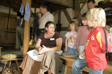 Kinder im Freilichtmuseum Eine Frau erklärt Kindern in rustikalem Ambiente die Zubereitung von traditionellen Knödeln.