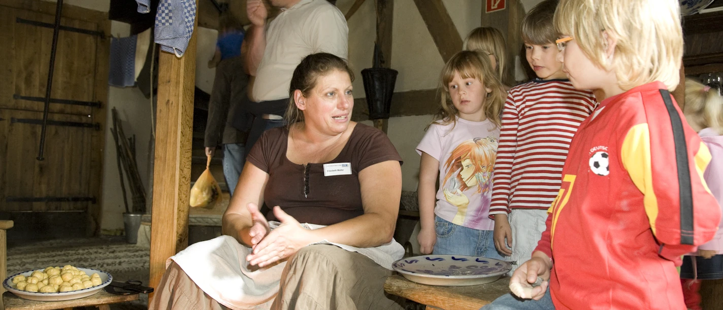 Kinder im Freilichtmuseum Eine Frau erklärt Kindern in rustikalem Ambiente die Zubereitung von traditionellen Knödeln.