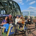 Rikolonia Fünf stilvoll gekleidete Personen sitzen in modernen Rikschas vor der Hohenzollernbrücke in Köln. Die Sonne scheint, der Himmel ist blau mit weißen Wolken. Five stylishly dressed people sit in modern rickshaws in front of the Hohenzollern Bridge in Cologne. The sun is shining, the sky is blue with white clouds.