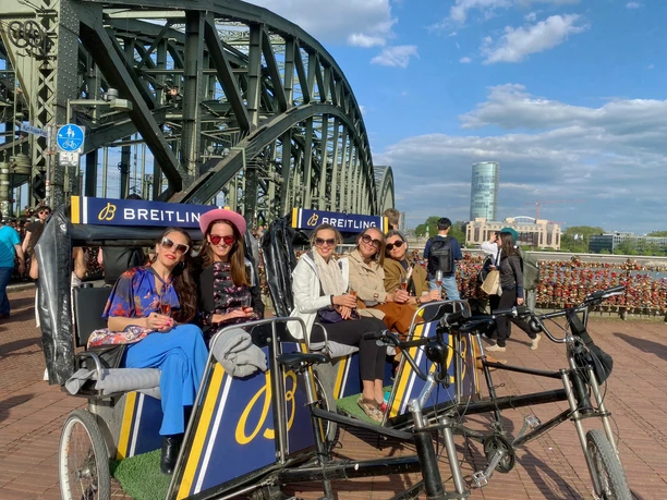 Rikolonia Fünf stilvoll gekleidete Personen sitzen in modernen Rikschas vor der Hohenzollernbrücke in Köln. Die Sonne scheint, der Himmel ist blau mit weißen Wolken.
