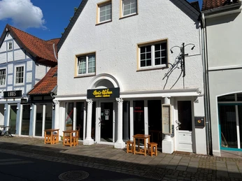 Hanis Kitchen Restaurantfassade mit weißen Säulen, Holztischen und schwarzem Schild, aufgenommen bei sonnigem Wetter.