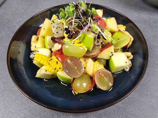 24 Yam Polamei.jpg Frischer Obstsalat mit Trauben, Äpfeln, Drachenfrucht, Mais und Microgreens, serviert auf einem Teller.
