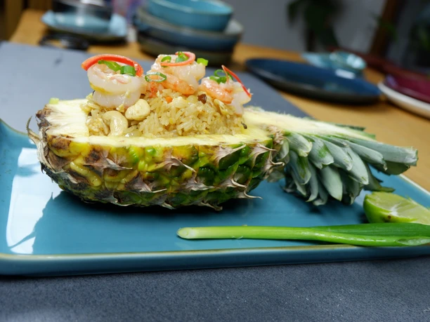 33 Khao Pad Saparrot.JPG Ananas gefüllt mit gebratenem Reis, Garnelen, Kräutern und Cashewnüssen auf einem blauen Teller serviert.
