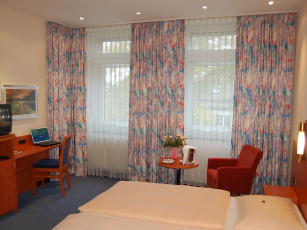 Hotelzimmer im Astoria Hotel in Ratingen Ein Hotelzimmer mit Doppelbett, Schreibtisch, Fernseher und Stuhl. Helle Vorhänge zieren die Fenster.