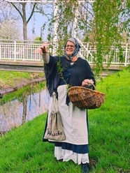 Auguste Eine Frau im historischen Kostüm, mit Kopftuch und Schürze, hält einen Korb. Sie steht lachend auf einer grünen Wiese neben dem Augustfehn-Kanal und einem Baum, im Hintergrund ist eine weiße Brücke zu sehen.