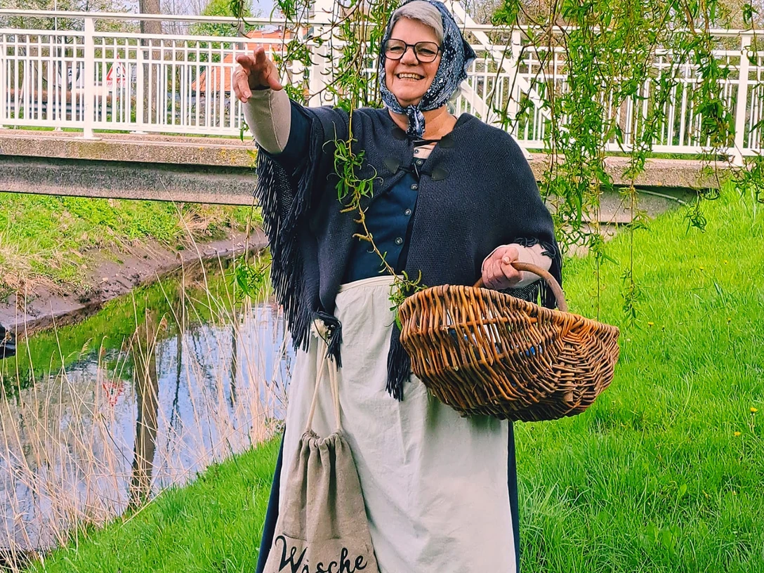 Auguste Eine Frau im historischen Kostüm, mit Kopftuch und Schürze, hält einen Korb. Sie steht lachend auf einer grünen Wiese neben dem Augustfehn-Kanal und einem Baum, im Hintergrund ist eine weiße Brücke zu sehen.