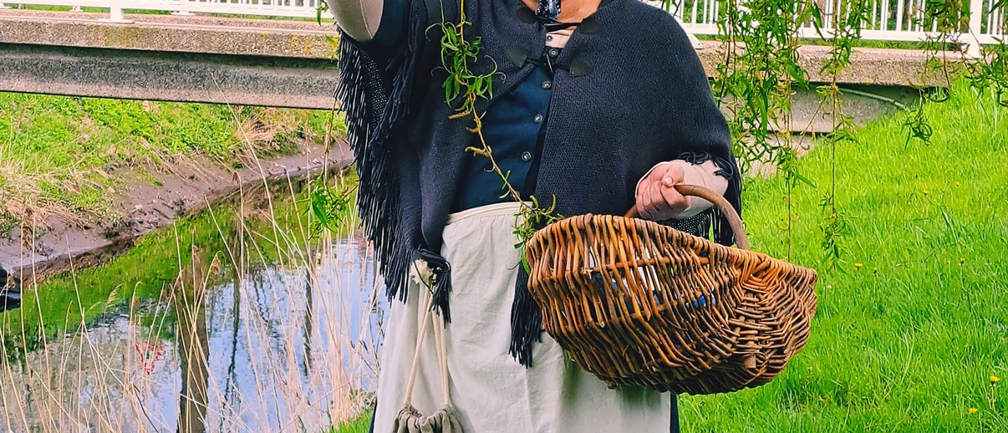 Auguste Eine Frau im historischen Kostüm, mit Kopftuch und Schürze, hält einen Korb. Sie steht lachend auf einer grünen Wiese neben dem Augustfehn-Kanal und einem Baum, im Hintergrund ist eine weiße Brücke zu sehen.