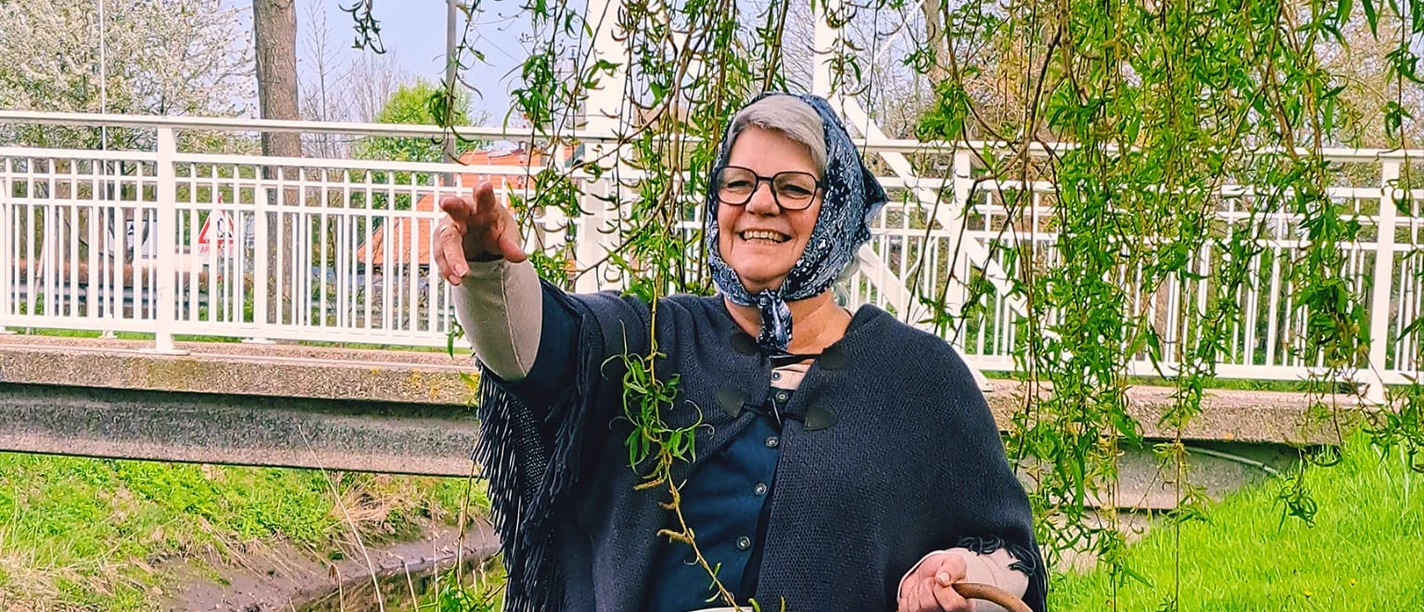 Auguste Eine Frau im historischen Kostüm, mit Kopftuch und Schürze, hält einen Korb. Sie steht lachend auf einer grünen Wiese neben dem Augustfehn-Kanal und einem Baum, im Hintergrund ist eine weiße Brücke zu sehen.