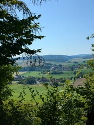 Burgweg Taubergsruh Blick durch Bäume auf grüne Felder und ein Dorf im Tal bei Einbeck unter blauem Himmel.