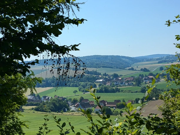 Burgweg Taubergsruh Blick durch Bäume auf grüne Felder und ein Dorf im Tal bei Einbeck unter blauem Himmel.