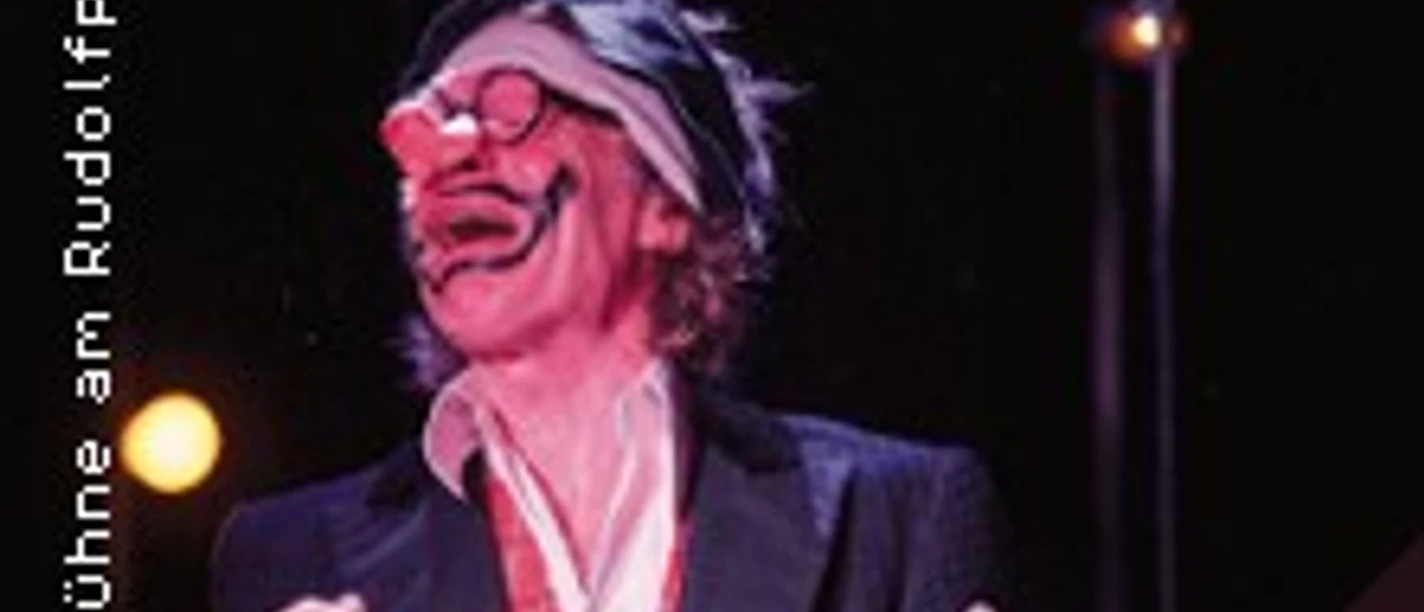 Karl Küpper in Clownskostüm mit roter Nase und Sonnebrille, lächelt bei Bühnenshow vor schwarzem Hintergrund.