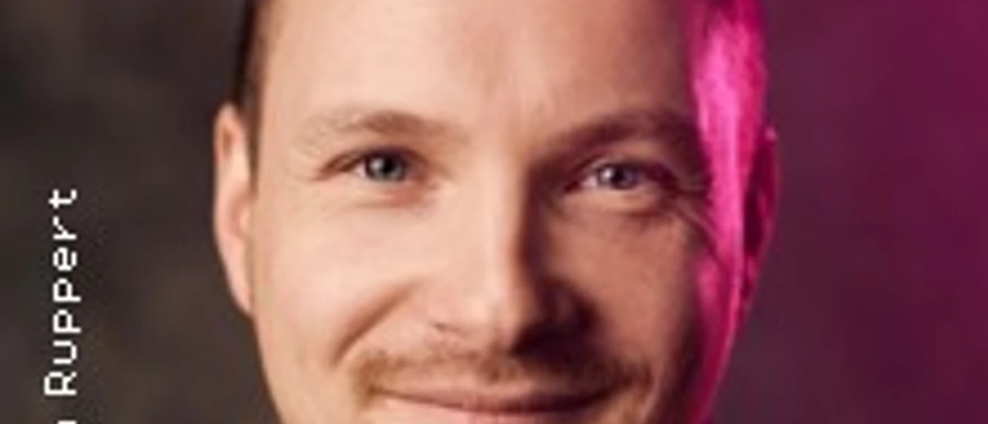 Porträt eines lächelnden Mannes mit Bart und kurzem Haar, von einem sanften Lichteffekt umgeben.