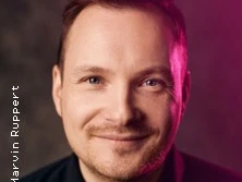 Porträt eines lächelnden Mannes mit Bart und kurzem Haar, von einem sanften Lichteffekt umgeben.