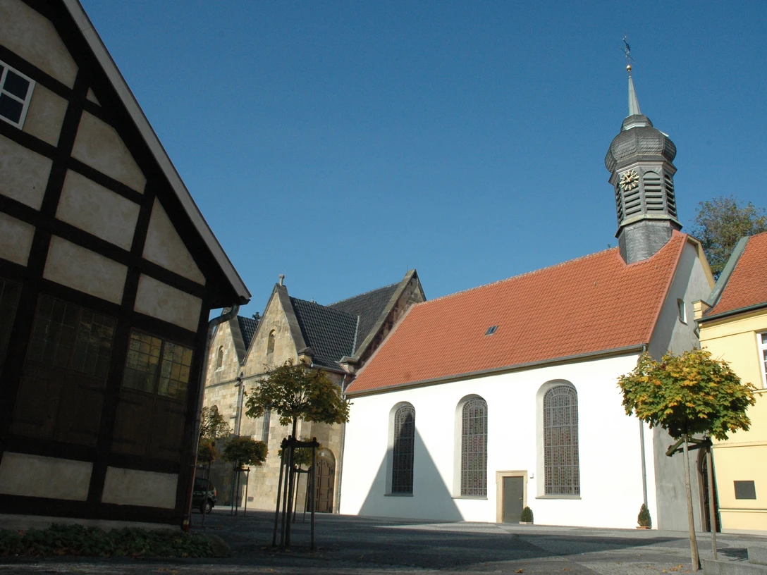 Fachwerkhaus, Kirche mit rotem Ziegeldach und Turm, umgeben von gepflastertem Platz und Bäumen.