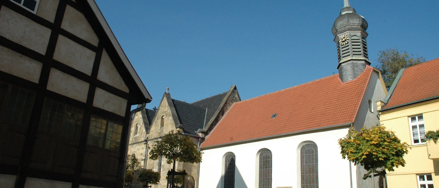 Fachwerkhaus, Kirche mit rotem Ziegeldach und Turm, umgeben von gepflastertem Platz und Bäumen.