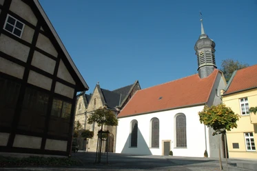 DSC_0545.JPG Fachwerkhaus, Kirche mit rotem Ziegeldach und Turm, umgeben von gepflastertem Platz und Bäumen.