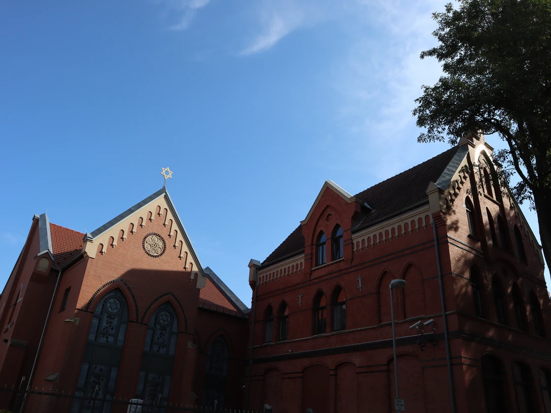 Synagoge (7).JPG Rote Backsteinsynagoge mit einem Davidstern auf dem Dach, umgeben von blauen Himmel und Bäumen.
