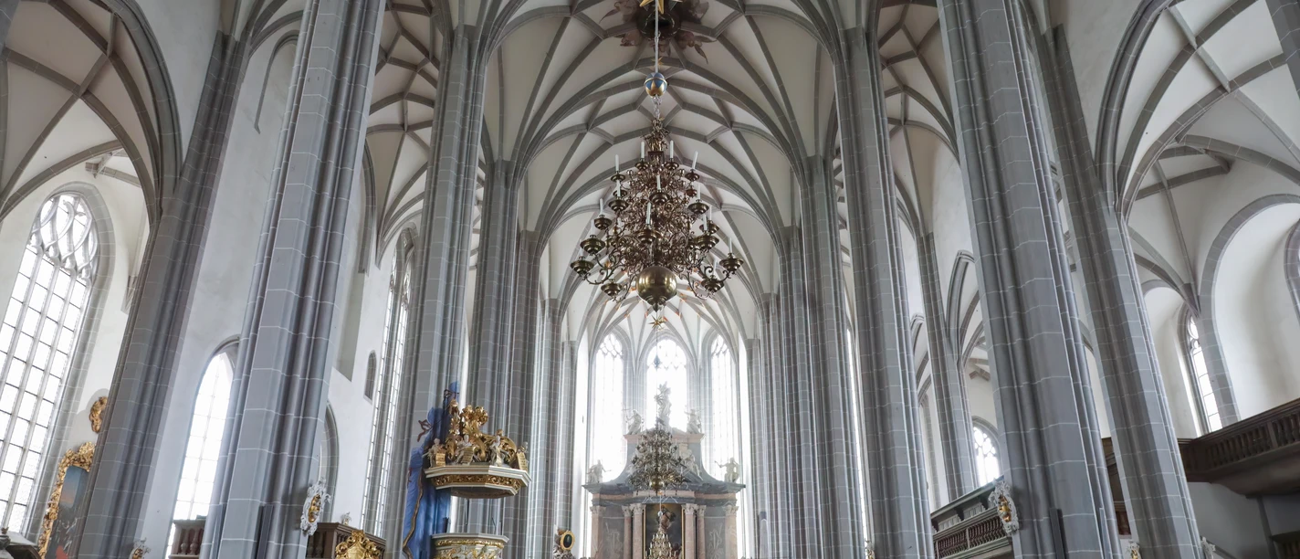 peterskirche-sonnenorgel-c-martin-doering-denoiseaI-low-light.jpg Innenansicht einer Kirche mit hohen Gewölben, kunstvollem Kronleuchter und hölzernem Gestühl.