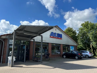 ALDI.jpg Ein ALDI-Supermarkt mit Backsteinfassade. Vor dem Gebäude stehen mehrere Autos. Der Himmel ist blau mit Wolken.