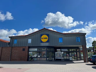 Lidl.jpg Ein Lidl Supermarkt mit grauem Dach und blauem Logo vor einem klaren Himmel mit Wolken.