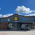 Lidl.jpg Ein Lidl Supermarkt mit grauem Dach und blauem Logo vor einem klaren Himmel mit Wolken.