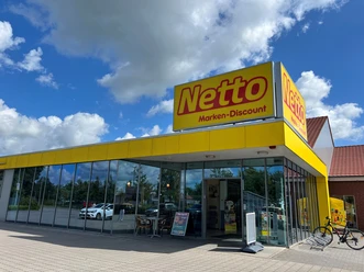 Netto.jpg Das Bild zeigt eine Netto-Filiale mit markant gelbem Schild und gläserner Fassade, umgeben von einem Parkplatz.