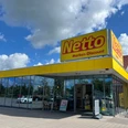 Netto.jpg Das Bild zeigt eine Netto-Filiale mit markant gelbem Schild und gläserner Fassade, umgeben von einem Parkplatz.