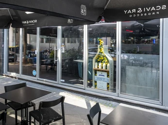 The Eat Ein Außenbereich mit schwarzen Tischen und Stühlen unter großen schwarzen Sonnenschirmen. Eine Glasfront zeigt das Innenleben des Gebäudes, und ein dekorativer Aufsteller in Form einer riesigen Limoncello-Flasche ist präsent.An outdoor seating area featuring black tables and chairs beneath large black sun umbrellas. A glass facade reveals the interior of the building, and a decorative stand shaped like a giant bottle of Limoncello is prominently displayed.