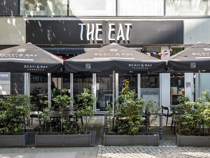The Eat Das Bild zeigt ein Restaurant namens "The Eat" mit mehreren Tischen unter schwarzen Sonnenschirmen. Der Eingangsbereich ist mit Pflanzen dekoriert.The image shows a restaurant named "The Eat," featuring several tables underneath black sun umbrellas. The entrance area is adorned with plants.