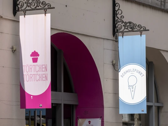 Toertchen Toertchen Zwei Schilder hängen über dem Eingang eines Gebäudes. Links ein Schild mit der Aufschrift "Törtchen Törtchen" auf rosa Hintergrund, rechts ein Schild für "Schmelzpunkt" mit einem Eis-Symbol auf blauem Hintergrund.