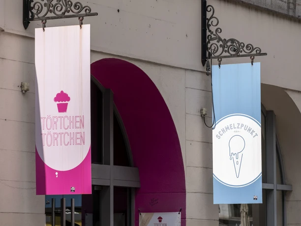 Toertchen Toertchen Zwei Schilder hängen über dem Eingang eines Gebäudes. Links ein Schild mit der Aufschrift "Törtchen Törtchen" auf rosa Hintergrund, rechts ein Schild für "Schmelzpunkt" mit einem Eis-Symbol auf blauem Hintergrund.