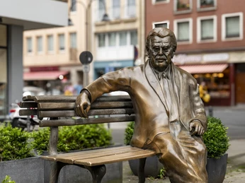 Willy Millowitsch memorial Bronzestatue von Willy Millowitsch auf einer Bank, umgeben von Büschen in urbaner Umgebung.Bronze statue of Willy Millowitsch on a bench, surrounded by bushes in an urban environment.