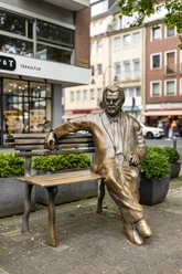Willy Millowitsch Denkmal Das Bild zeigt eine bronzene Statue von Willy Millowitsch, die auf einer Parkbank sitzend dargestellt ist. Die Statue befindet sich in einer urbanen Umgebung, flankiert von Blumenbeeten und Gebäuden.