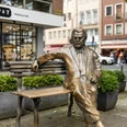 Willy Millowitsch Denkmal Das Bild zeigt eine bronzene Statue von Willy Millowitsch, die auf einer Parkbank sitzend dargestellt ist. Die Statue befindet sich in einer urbanen Umgebung, flankiert von Blumenbeeten und Gebäuden.The picture shows a bronze statue of Willy Millowitsch sitting on a park bench. The statue is located in an urban setting, flanked by flowerbeds and buildings.