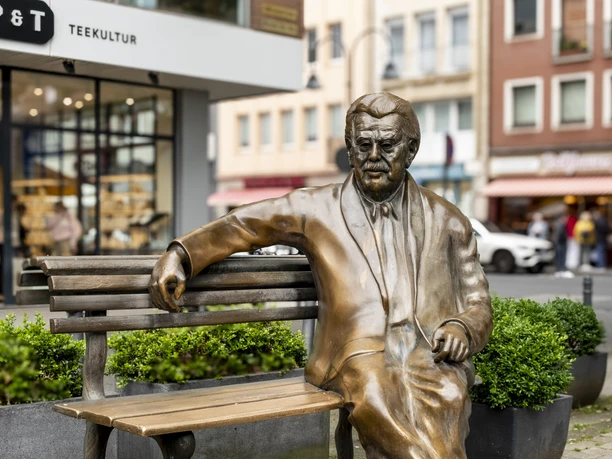 Willy Millowitsch Denkmal Das Bild zeigt eine bronzene Statue von Willy Millowitsch, die auf einer Parkbank sitzend dargestellt ist. Die Statue befindet sich in einer urbanen Umgebung, flankiert von Blumenbeeten und Gebäuden.