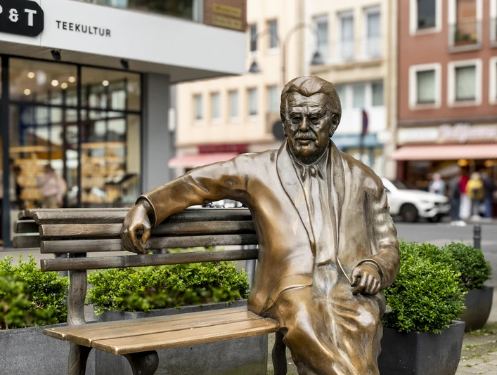 Willy Millowitsch memorial Das Bild zeigt eine bronzene Statue von Willy Millowitsch, die auf einer Parkbank sitzend dargestellt ist. Die Statue befindet sich in einer urbanen Umgebung, flankiert von Blumenbeeten und Gebäuden.The picture shows a bronze statue of Willy Millowitsch sitting on a park bench. The statue is located in an urban setting, flanked by flowerbeds and buildings.