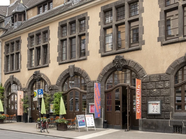 Comedia Das Bild zeigt die Fassade des historischen Gebäudes der Comedia in Köln, ein kulturelles Zentrum mit großen Sprossenfenstern und dekorativen Steinornamenten. Vor der Gebäudezeile laden Schirme und Sitzmöbel unter freiem Himmel zum Verweilen ein.