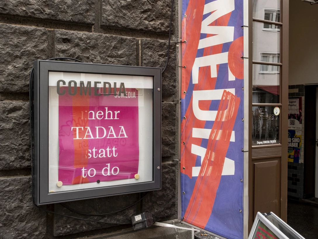 Comedia Der Eingang der Comedia-Theater in Köln zeigt bunte Fahnen und ein beleuchtetes Plakat, das vorbeigehende Menschen dazu einlädt, die lebendige Kulturszene zu entdecken.The entrance to the Comedia theaters in Cologne displays colorful flags and an illuminated poster inviting passers-by to discover the lively cultural scene.