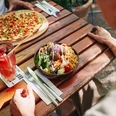 Stadtgarten Ein bunt gedeckter Holztisch zeigt einen sommerlichen Tisch im Freien mit einem frischen Salat und Pizza.A colorfully set wooden table showcases a summery outdoor dining scene, featuring a fresh salad and pizza.