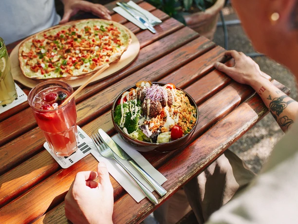 Stadtgarten Ein bunt gedeckter Holztisch zeigt einen sommerlichen Tisch im Freien mit einem frischen Salat und Pizza.