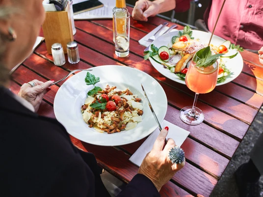 Stadtgarten Eine stilvolle Tischszene im Freien. Im Vordergrund ein Teller mit Pasta garnierte Kirschtomaten und Kräutern.A stylish outdoor table setting. In the foreground, a plate of pasta garnished with cherry tomatoes and herbs.