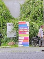 Cologne Book Festival A colorful advertising banner for the Cologne Book Festival stands in front of an overgrown wall, with a bicycle next to it.