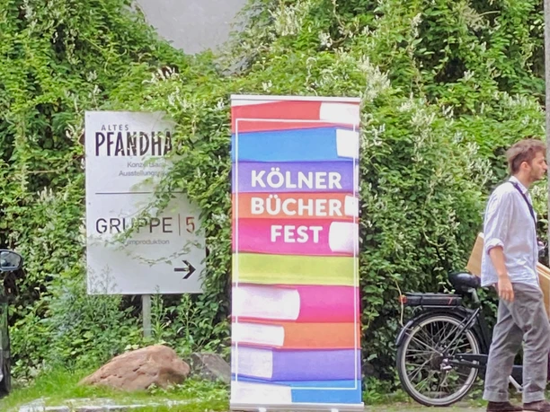 Cologne Book Festival A colorful advertising banner for the Cologne Book Festival stands in front of an overgrown wall, with a bicycle next to it.