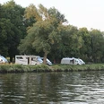 Wohnmobile parken entlang eines Flusses, von Bäumen gesäumt; im Hintergrund eine Brücke sichtbar.