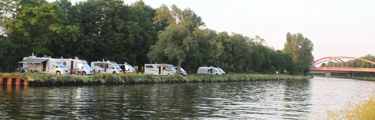 Wohnmobile02.jpg Wohnmobile parken entlang eines Flusses, von Bäumen gesäumt; im Hintergrund eine Brücke sichtbar.